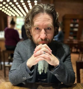 Photo of the author in a blazer and t-shirt facing the camera with fingers interlocked.